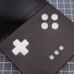 May include: A black handheld gaming console with a white cross-shaped button and four white round buttons. The console has a black screen and the text "Analogue Pocket" is visible on the front.