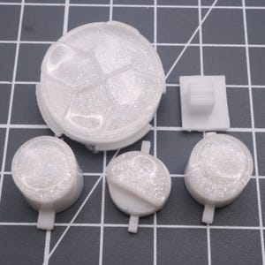 May include: A set of five white plastic buttons with a sparkly finish. The buttons are round and have a raised design. The buttons are perfect for adding a touch of sparkle to any project.