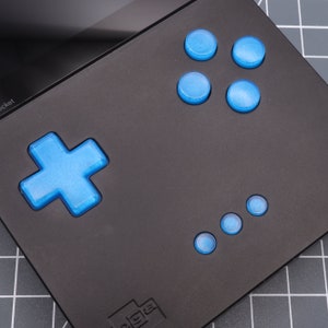 Op de afbeelding: Een zwart draagbaar gameapparaat met een blauwe kruisvormige knop, vier blauwe ronde knoppen en vier kleinere blauwe ronde knoppen. Het apparaat heeft een klein scherm en de tekst "Pocket" is zichtbaar in de linkerbovenhoek.