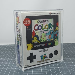 Può includere: Una custodia in plastica trasparente con una console per videogiochi portatile Game Boy Color bianca all'interno. La console ha uno schermo nero con un'immagine colorata di personaggi Pokémon. Il testo "GAME BOY COLOR" è stampato sulla console in bianco e nero. Il testo "Nintendo" è stampato sulla console in bianco. Il testo "CGB-001" è stampato in nero nella parte inferiore della console.
