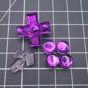 May include: A set of purple replacement buttons for a video game controller. The set includes a directional pad, four face buttons, and four clear plastic button stems.