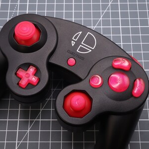 May include: A black GameCube controller with red buttons. The controller features a white Nintendo logo and the words "Start/Pause" printed on it.