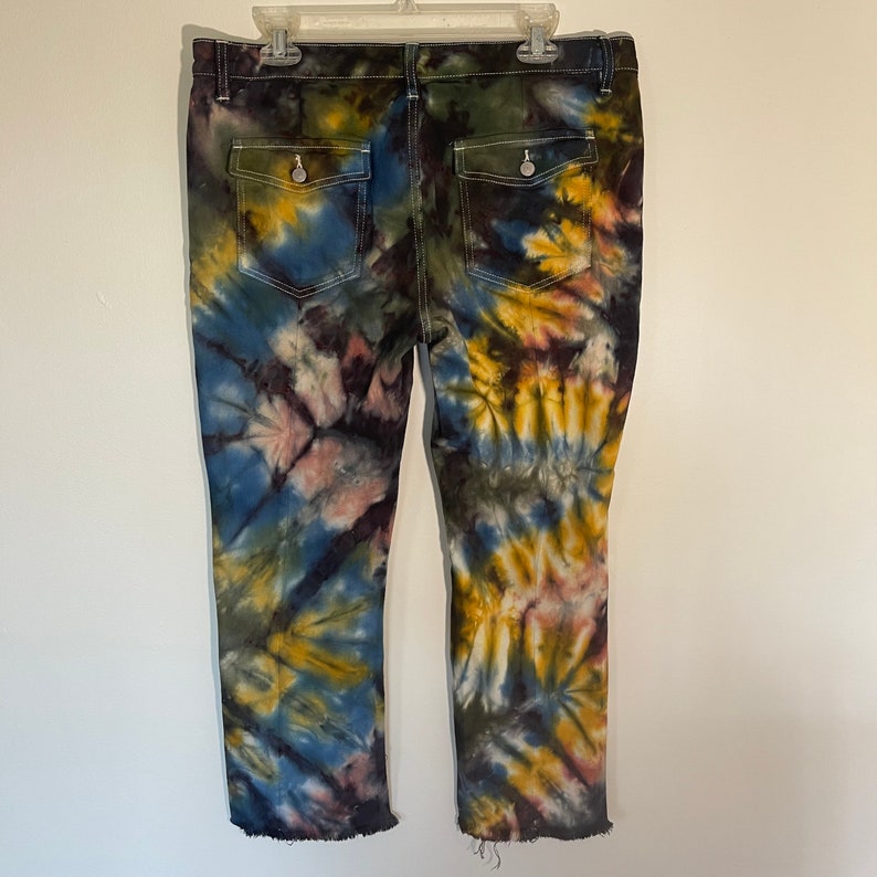 May include: Tie-dye jeans with a unique pattern in shades of blue, yellow, pink, and green. The jeans have two back pockets with button closures and a raw hem. The jeans are hanging on a white plastic hanger.