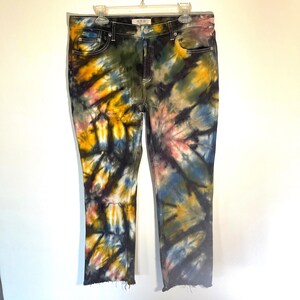 May include: Tie-dye jeans in a variety of colors, including yellow, blue, pink, and green. The jeans have a classic five-pocket design, a button and zipper closure, and a frayed hem. The jeans are hanging on a white hanger.