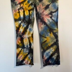 May include: Tie-dye jeans in a unique pattern featuring shades of yellow, blue, pink, and black. The jeans have a straight leg cut with a raw hem. The stitching is white, and the jeans are displayed against a white background.