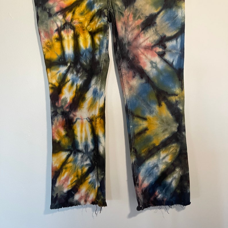 May include: Tie-dye jeans in a unique pattern featuring shades of yellow, blue, pink, and black. The jeans have a straight leg cut with a raw hem. The stitching is white, and the jeans are displayed against a white background.