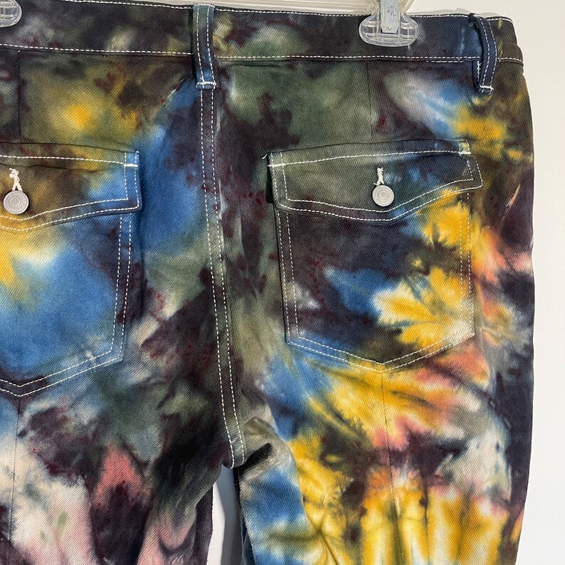 May include: Close-up of the back of tie-dye jeans. The jeans feature a vibrant mix of colors including blue, yellow, green, and maroon. White stitching outlines the pockets and seams. The jeans have two back pockets and a button closure.