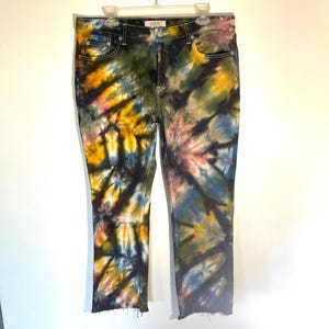 May include: Tie-dye jeans in a straight-leg style. The denim is dyed with a spiral pattern in shades of yellow, blue, pink, and black. The jeans have a classic five-pocket design and a raw hem. The jeans are hanging on a white hanger.