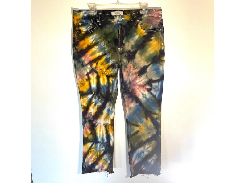 May include: Tie-dye jeans in a straight-leg style. The denim is dyed with a spiral pattern in shades of yellow, blue, pink, and black. The jeans have a classic five-pocket design and a raw hem. The jeans are hanging on a white hanger.