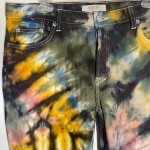 May include: Close-up of tie-dye jeans with a mix of colors including yellow, blue, pink, and black. The jeans have white stitching and silver rivets. The tag reads "WE THE FREE".