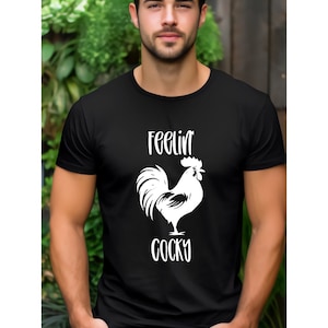 Puede incluir: Camiseta negra de cuello redondo con un gráfico de gallo blanco y las palabras "Feelin' Cocky". La camiseta está hecha de un material suave y es adecuada para ropa casual.
