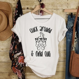 Puede incluir: Camiseta blanca con un gráfico negro de un pollo con gafas y el texto "Cluck around & find out".