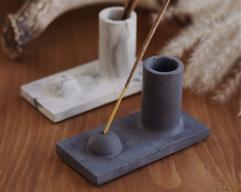 May include: Two concrete incense holders, one white and one gray. The gray holder has a round indentation in the center and a cylindrical opening for incense sticks. The white holder has a rectangular base and a cylindrical opening for incense sticks.