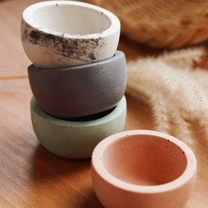 May include: Four small, round concrete bowls in a stack. The colours are white with black marbling, grey, green, and peach. They sit on a wooden surface. A woven basket and dried plants are in the background. Measurements are in centimetres.