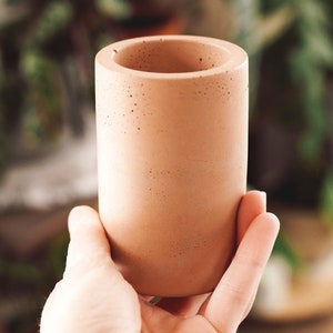May include: A light brown concrete planter with a smooth, slightly textured surface. The planter is cylindrical in shape and has a wide opening at the top.