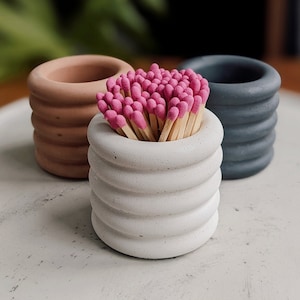 May include: Three concrete matchstick holders in different colors: brown, white, and gray. The white holder has pink matchsticks inside.