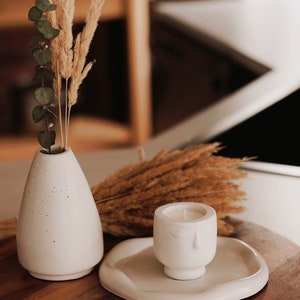 Pu&ograve; includere: Un vaso in ceramica bianca con una superficie testurizzata contiene erba della pampa secca e rami di eucalipto. Un portacandele in ceramica bianca con un design a forma di viso si trova su un vassoio in ceramica bianca su una superficie in legno.