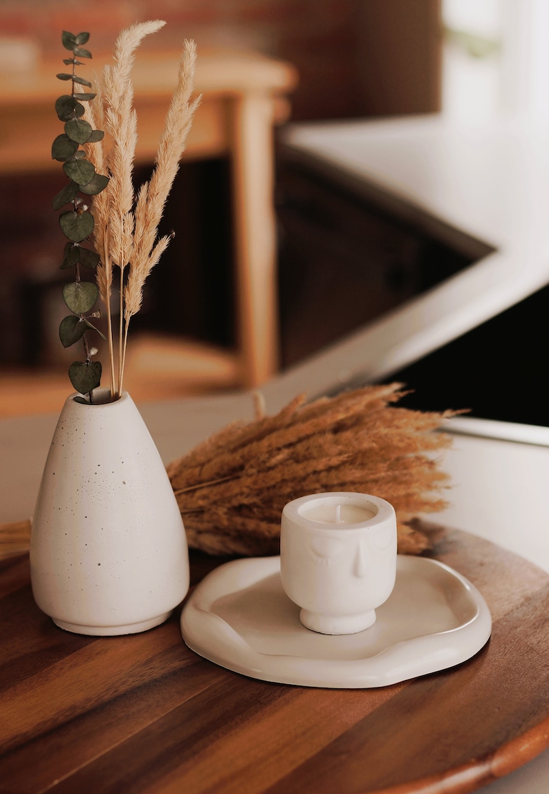 Pu&ograve; includere: Un vaso in ceramica bianca con una superficie testurizzata contiene erba della pampa secca e rami di eucalipto. Un portacandele in ceramica bianca con un design a forma di viso si trova su un vassoio in ceramica bianca su una superficie in legno.