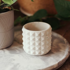 May include: A white concrete planter with a geometric pattern. The planter is sitting on a grey and white marble surface.