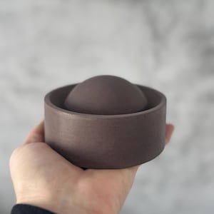 Peut inclure: Un récipient en céramique brun foncé avec un couvercle arrondi, tenu dans une main. Le récipient a une base circulaire et une finition lisse et mate. Le design est minimaliste et moderne, adapté aux petits objets ou à un usage décoratif.