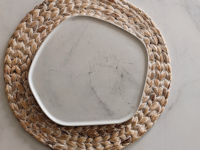 Puede incluir: Un plato de cer&aacute;mica blanco con una forma ligeramente irregular, apoyado en un mantel individual de paja trenzada.