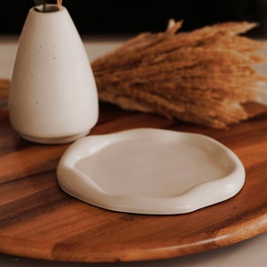 Pu&ograve; includere: Un vaso di ceramica bianca con una piccola pianta all'interno &egrave; posto su un giradischi di legno con un piatto di ceramica bianca. Il giradischi &egrave; circondato da steli di grano secchi.