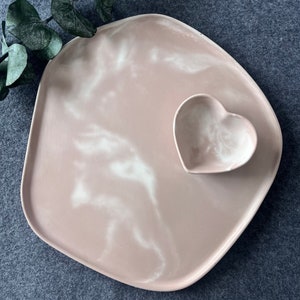 Puede incluir: Una bandeja de servir de m&aacute;rmol rosa y blanco con un plato en forma de coraz&oacute;n. La bandeja tiene una forma &uacute;nica e irregular y es perfecta para servir aperitivos o aperitivos.