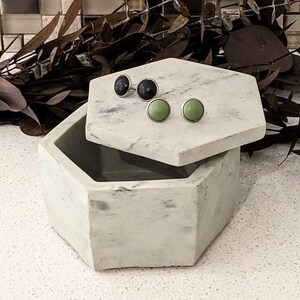 May include: A white marble hexagonal box with a lid, open to reveal two pairs of earrings. One pair is black and the other is green.