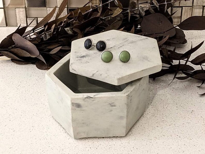 May include: A white marble hexagonal box with a lid, open to reveal two pairs of earrings. One pair is black and the other is green.