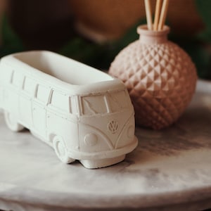 May include: White ceramic planter shaped like a vintage Volkswagen bus. The planter has a small opening on top for planting. The planter is sitting on a grey marble tray with a pink ceramic diffuser in the background.