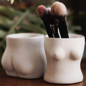 May include: Two white ceramic containers shaped like a human torso and buttocks, holding a set of makeup brushes with pink, brown, and black bristles.
