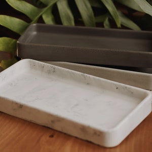 May include: Two rectangular trays, one white with a marbled pattern and one grey, are stacked on top of each other. The trays are made of concrete and have a rough texture.