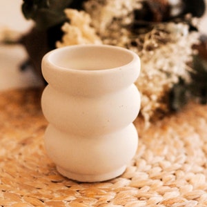 Peut inclure: Un vase en céramique blanche avec un design ondulé et texturé. Le vase est posé sur un tapis de paille tissé.