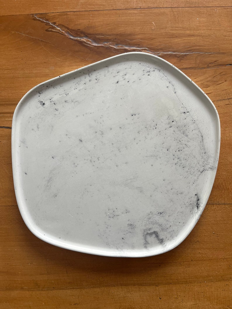 Puede incluir: Un plato de cer&aacute;mica blanco con una forma ligeramente irregular y una superficie texturizada moteada. El plato tiene un aspecto ligeramente desgastado con motas negras en toda la superficie.
