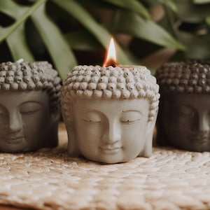 May include: Three grey concrete Buddha head candle holders, one with a lit candle, on a woven straw mat.