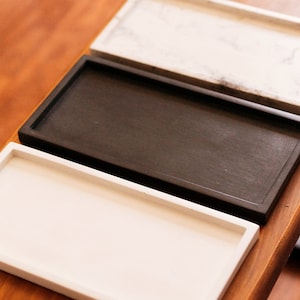 May include: Three rectangular trays, one white, one black, and one white with grey marbling, are stacked on top of each other on a wooden surface.