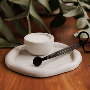 Pu&ograve; includere: Un portacandele in ceramica bianca con una candela bianca all'interno, uno spegnicandele in metallo nero e un vassoio in ceramica bianca.