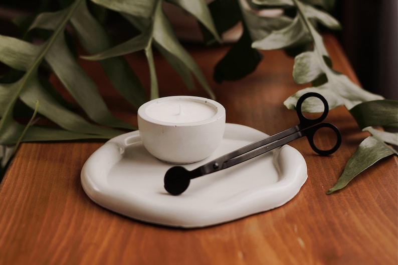 Pu&ograve; includere: Un portacandele in ceramica bianca con una candela bianca all'interno, uno spegnicandele in metallo nero e un vassoio in ceramica bianca.