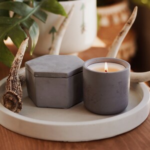 May include: A gray concrete tray with a round rim holds a gray concrete candle and a gray concrete box. The candle is lit and the tray is decorated with two antlers.