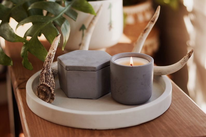 May include: A gray concrete tray with a round rim holds a gray concrete candle and a gray concrete box. The candle is lit and the tray is decorated with two antlers.