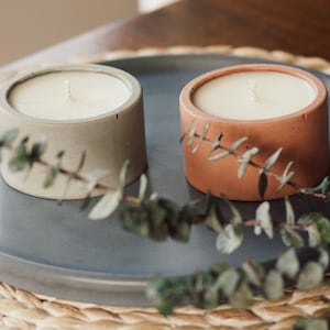 Peut inclure: Deux bougeoirs en béton avec une bougie blanche dans chacun. Les bougeoirs sont sur un plateau gris avec un tapis tissé en dessous. Une branche d'eucalyptus se trouve devant les bougies.