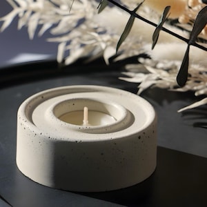 May include: A white concrete candle holder with a circular design and a small white candle in the centre. The holder is sitting on a black surface.