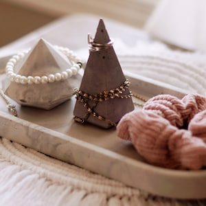 May include: A grey concrete tray with a diamond-shaped and a pyramid-shaped ring holder. The tray also has a pearl necklace and a gold chain. A pink scrunchie is on the tray.