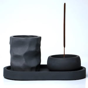 May include: A grey concrete incense holder with a textured surface. The holder has a small bowl for incense and a larger bowl for a plant or other decorative item. An incense stick is placed in the bowl.