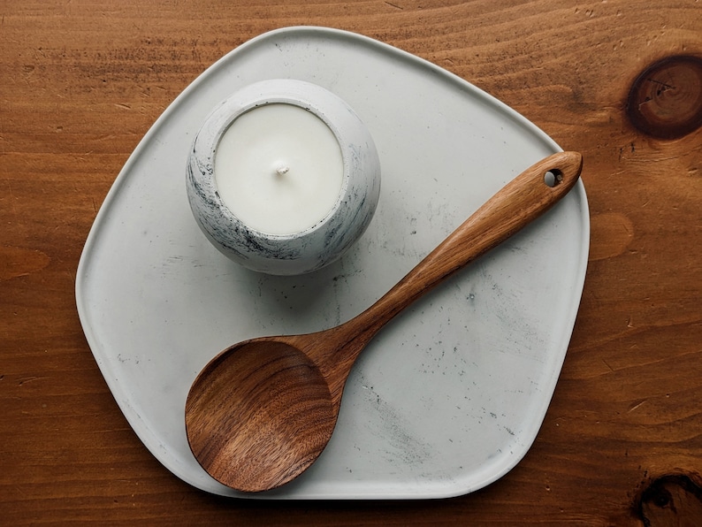 Puede incluir: Una bandeja de cer&aacute;mica blanca con una cuchara de madera y una vela blanca con un acabado moteado gris.