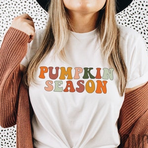 May include: A white t-shirt with the text "Pumpkin Season" in colorful, retro-style letters. The letters are in shades of orange, green, yellow, and pink.