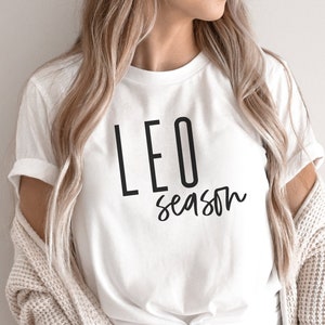 May include: White t-shirt with black text that reads "LEO season". The person wearing the shirt is also wearing a light brown cardigan and blue jeans.