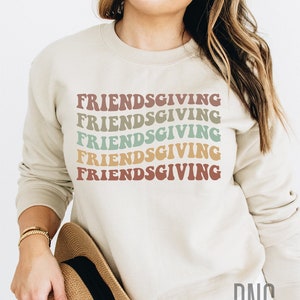 May include: A beige sweatshirt with the text "Friendsgiving" repeated five times in a retro font with different colors. The text is stacked vertically down the center of the sweatshirt.