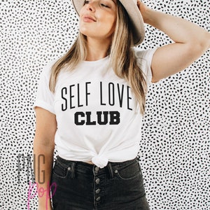 Puede incluir: Una camiseta blanca con texto negro que dice "SELF LOVE CLUB". La camiseta est&aacute; anudada a la cintura y la modelo lleva pantalones vaqueros negros y un sombrero beige.