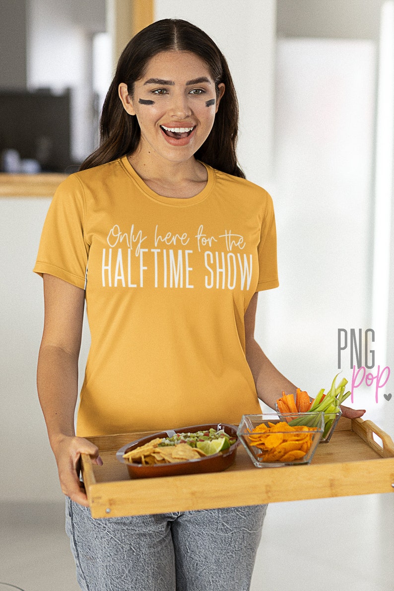 K&ouml;nnte beinhalten: Eine Frau tr&auml;gt ein gelbes T-Shirt mit dem Text "Only here for the HALFTIME SHOW" und h&auml;lt ein Holztablett mit einer Sch&uuml;ssel Guacamole und Chips, einer Sch&uuml;ssel Chips und einer kleinen Sch&uuml;ssel Gem&uuml;se.